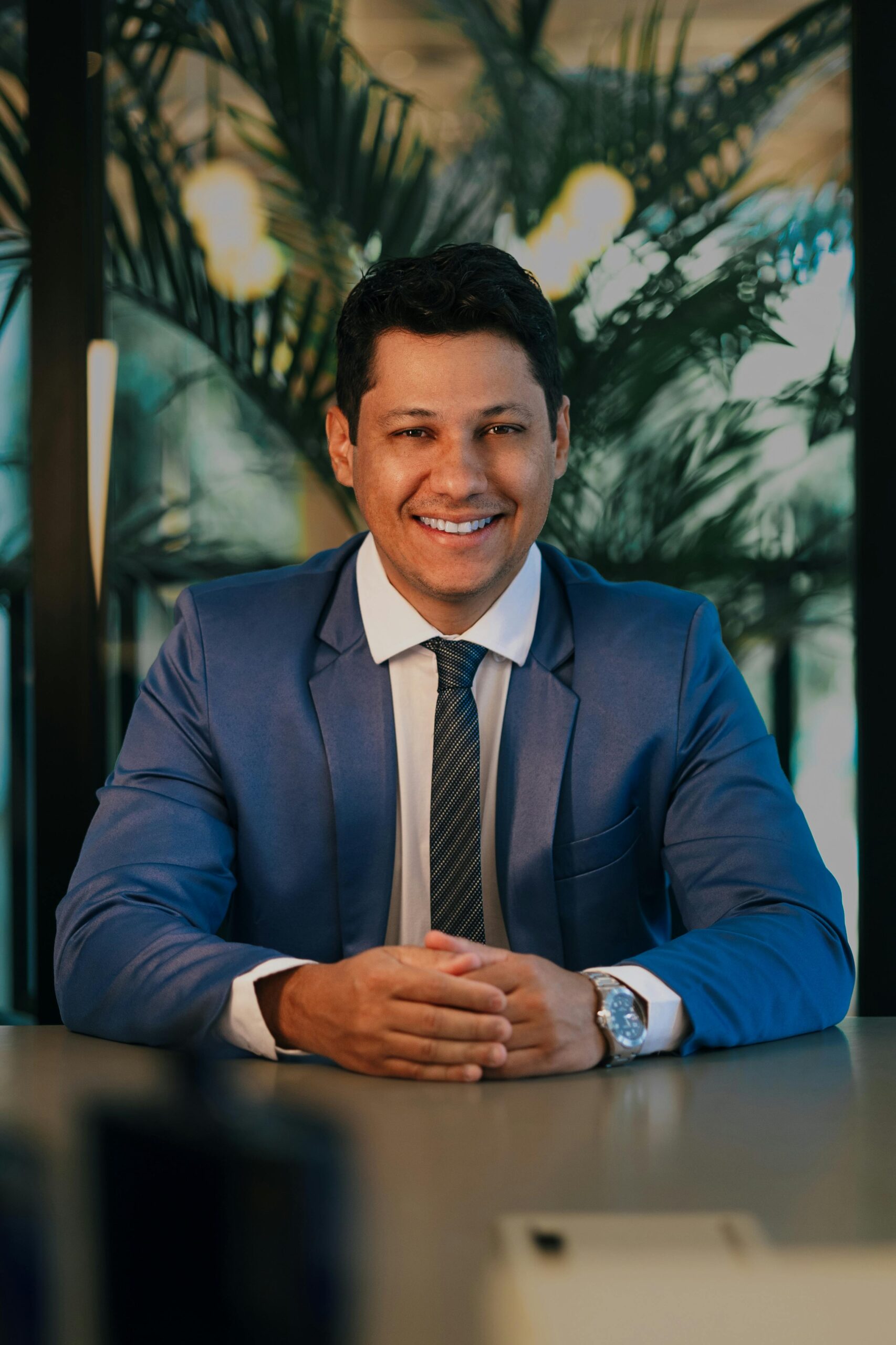 Confident businessman in a blue suit smiling at a desk in a modern office.